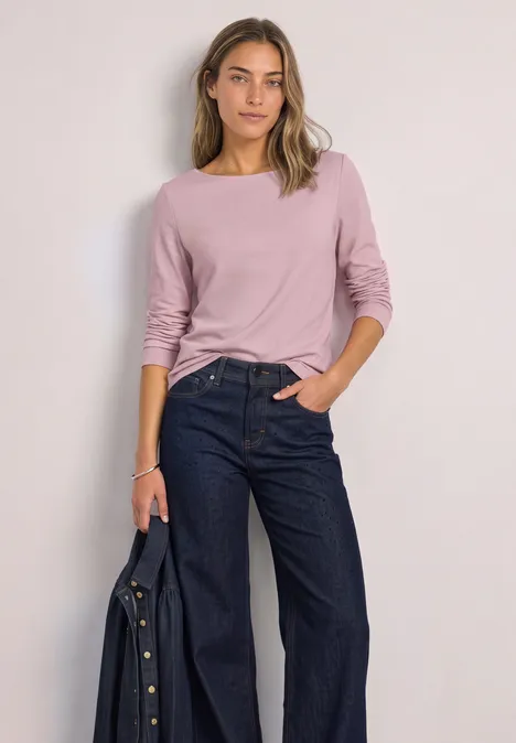 Basic shirt met boothals quiet rose Frau in lockerem rosa Langarmshirt und weitem Jeans, hält eine Jacke in der Hand.