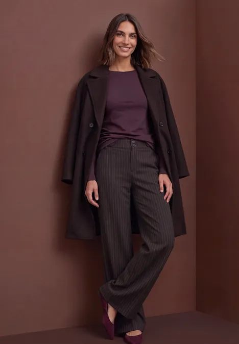 Vrouw voor een bruine achtergrond, met een bruine mantel, bordeaux top en wijde broek met fijne strepen.