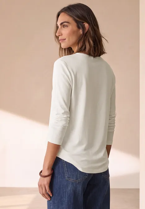 Frau in weißem Langarmshirt und Jeans, die lässig zur Seite blickt.