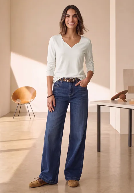 Frau in hellem Raum trägt weiße V-Ausschnitt-Bluse, hohe blaue Weitbein-Jeans und braune Loafers.