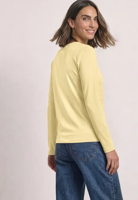 Frau in gelbem Langarmshirt und blauen Jeans, die über die Schulter lächelnd zurückblickt.