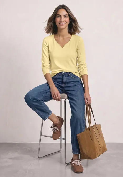 Frau in gelbem Oberteil und blauen Jeans sitzt auf einem Hocker, lächelt und hält eine braune Tasche.