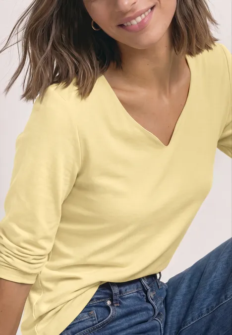 Frau mit gelbem V-Ausschnitt-Shirt und blauer Jeans, sitzt lächelnd.
