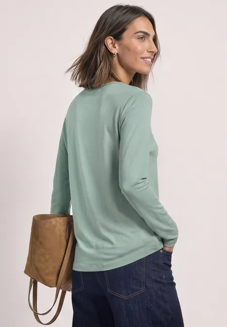 Frau mit kurzem Braunhaar schaut rückwärts, trägt grünes Langarmshirt, dunkle Jeans und eine braune Tasche.