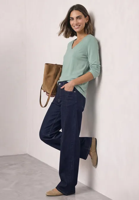 Eine Frau lehnt an der Wand, trägt hellgrünen Pullover, weite Jeans und braune Stiefeletten.