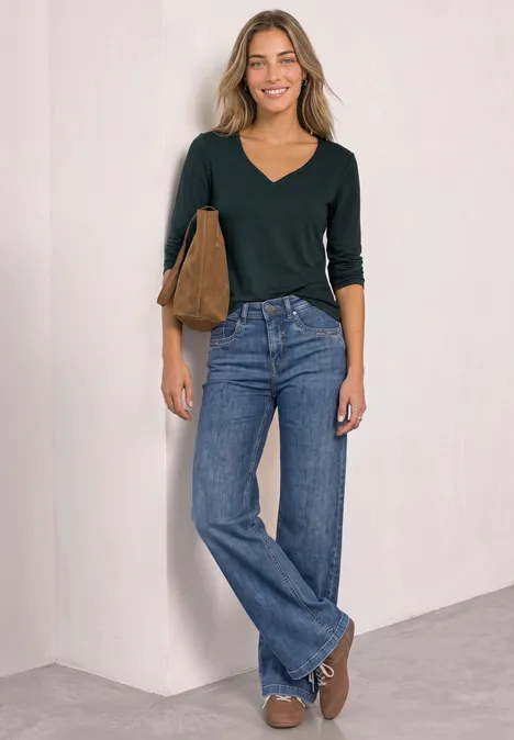 Eine lächelnde Frau in grüner V-Neck-Bluse, weiten Jeans und brauner Tasche vor neutralem Hintergrund.