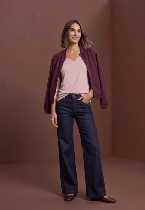Een glimlachende vrouw in een paarse bomberjack, roze top en wijde, taillehoge jeans voor een bruine achtergrond.