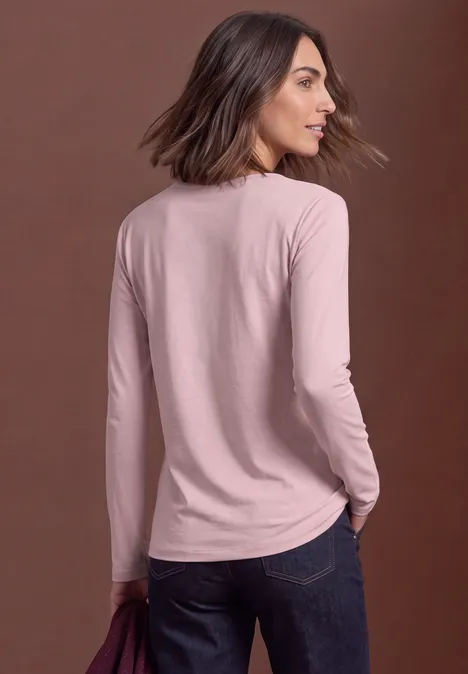 Vrouw in een roze longsleeve en spijkerbroek, licht van achteren, kijkt over haar schouder tegen een neutrale achtergrond.