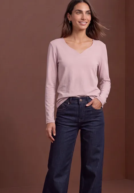 Vrouw met roze V-hals shirt en wijd donkerblauwe spijkerbroek voor een bruine achtergrond.