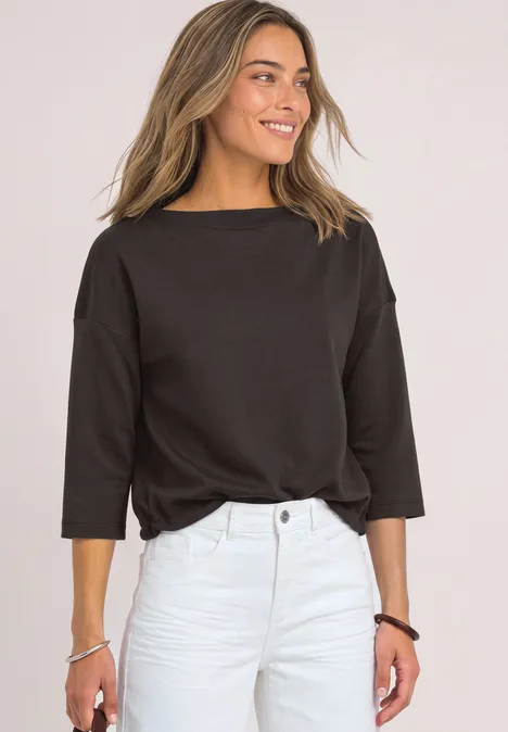 Vrouw glimlacht in zwart shirt met 3/4-mouwen en lichte broek voor een lichte achtergrond.