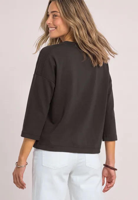 Vrouw in zwarte trui en witte broek kijkt lachend over haar schouder.