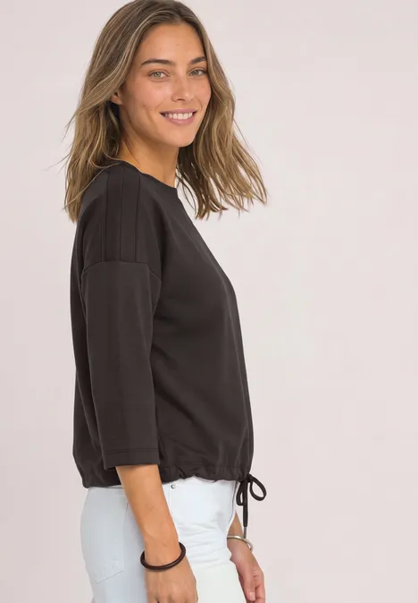 Een lachende vrouw in profiel draagt een zwarte 3/4-mouw top met strikkoord en witte broek tegen een lichte achtergrond.