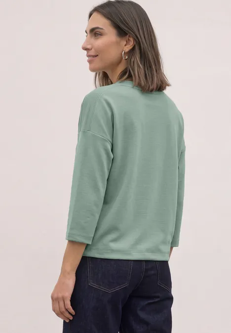 Vrouw in profiel met losse groene 3/4-mouw top en donkere spijkerbroek voor een neutrale achtergrond.
