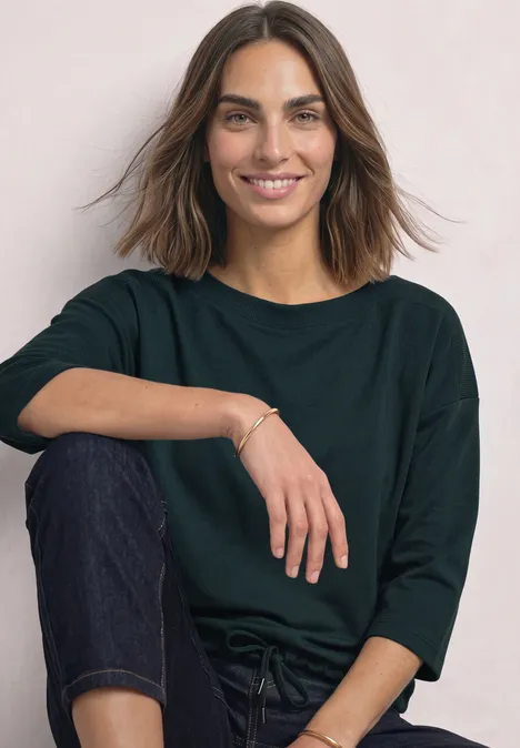 Een glimlachende vrouw in een groen shirt en spijkerbroek zit ontspannen voor een lichte achtergrond.