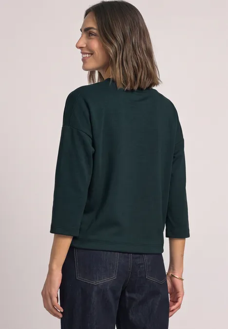 Vrouw van achteren met een donkergroene trui met driekwartmouwen en spijkerbroek voor een lichte achtergrond.