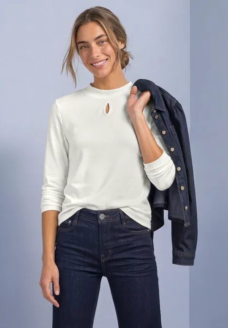 Lächelnde Frau in weißem Langarmshirt und dunklen Jeans mit Jacke über der Schulter.