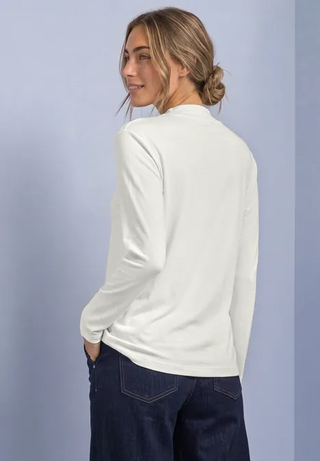 Frau steht mit Blick über die Schulter, trägt weißen Langarmshirt und dunkle Jeans vor neutralem Hintergrund