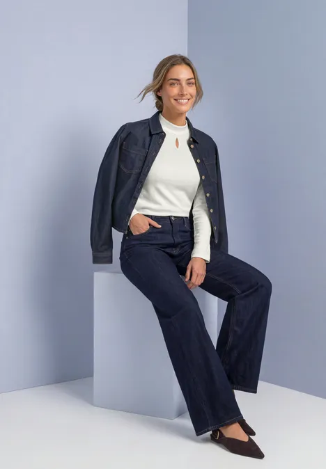 Frau in dunklem Jeans-Outfit und weißem Top, lächelnd auf einem hellen Würfel sitzend vor neutralem Hintergrund.