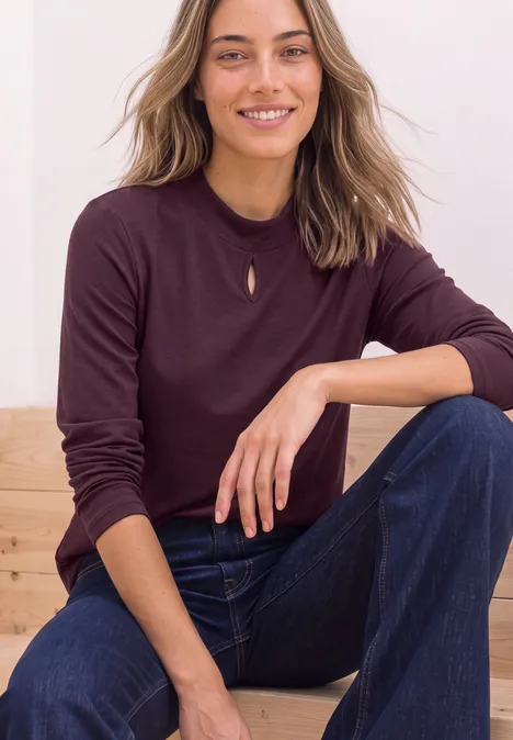 Frau sitzt lächelnd auf einer Bank, trägt einen auberginefarbenen Pullover mit kleinem Ausschnitt und blaue Jeans.