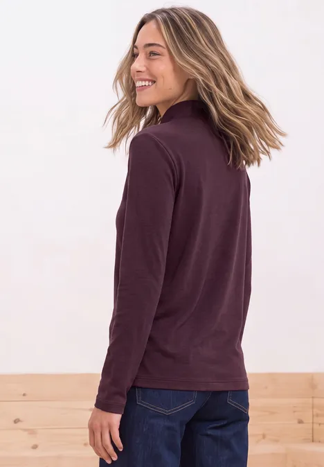 Eine seitlich lächelnde Frau in burgunderfarbenem Pullover und Jeans mit weichen blonden Haaren.