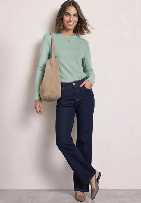 Frau in grünem Langarmshirt, dunklen Jeans, brauner Tasche auf der Schulter, lächelt vor hellem Hintergrund.