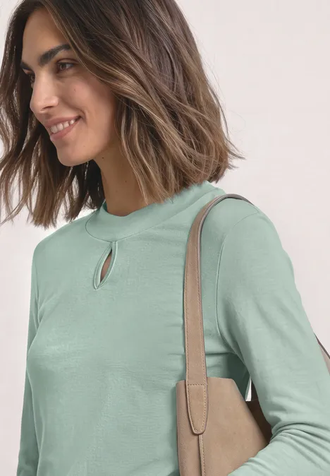 Frau mit schulterlangem braunem Haar trägt grünes Langarmshirt mit Schlitz am Hals und beiger Schultertasche