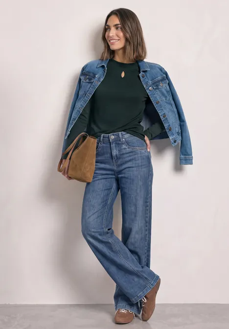 Vrouw met spijkerjasje over de schouders, groen truitje, wijde jeans, bruine laarsjes en schoudertas.