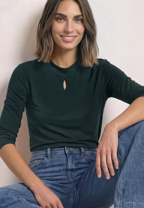 Een glimlachende vrouw zit ontspannen, gekleed in een donkergroen shirt met lange mouwen en een blauwe jeans.