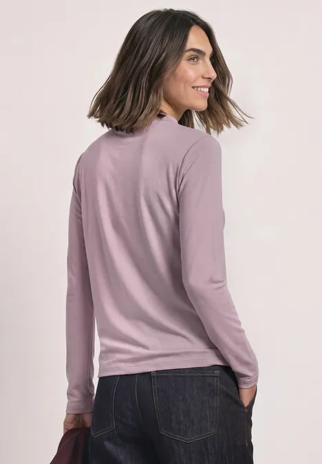 Frau mit langärmligem rosafarbenem Pullover und Jeans, schaut über die Schulter und lächelt.