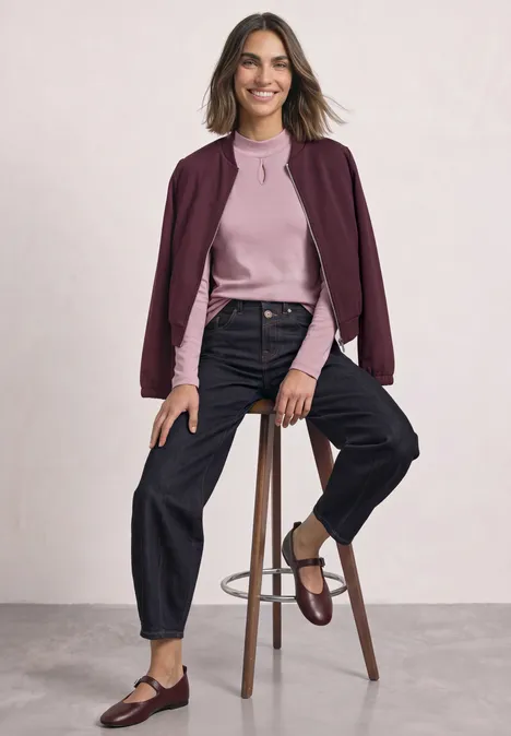 Frau sitzt auf Hocker in rosa Pullover, bordeaux Jäckchen, schwarzen Jeans und braunen Loafers.