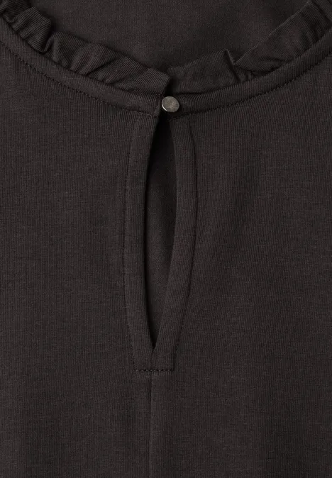 Schwarzes Shirt, Rundhalsausschnitt mit Rüsche, kleiner Schlitz mit Knopf und Bindeband.