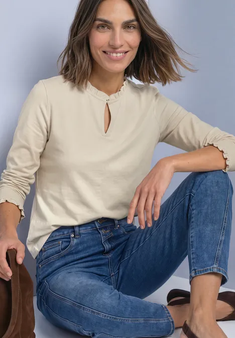 Basic Shirt mit Rüschen und Cut-Out-Detail pure beige Frau kniet lässig, trägt beiges Langarmshirt mit Rüschen und blaue Jeans.