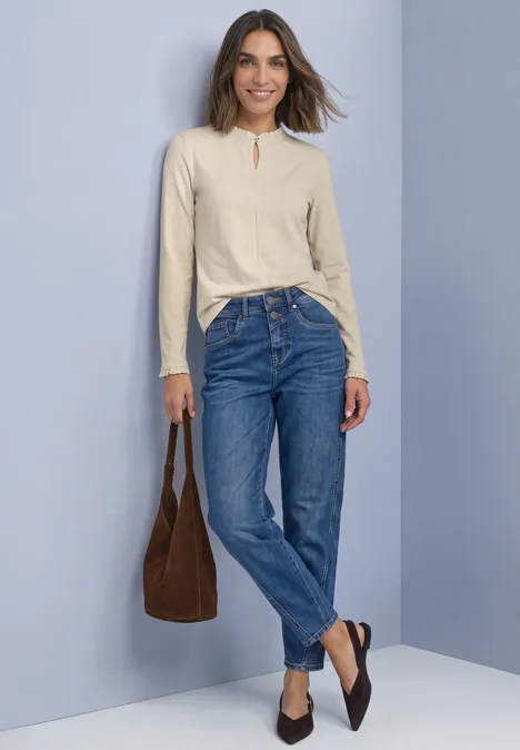 Basic Shirt mit Rüschen und Cut-Out-Detail pure beige Frau in beiger Bluse, blauen Jeans, brauner Tasche und dunklen Loafers vor hellgrauer Wand, lächelnd.