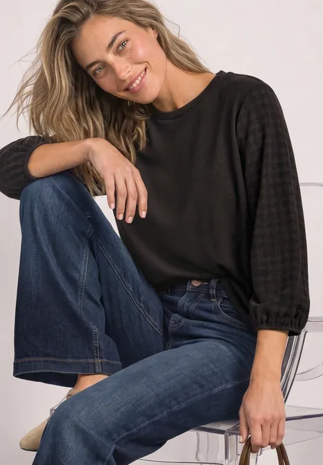 Shirt mit Rundhals und Strukturärmeln black coffee Eine lächelnde Frau sitzt entspannt auf einem Stuhl, trägt schwarzen Pullover und blaue Jeans.