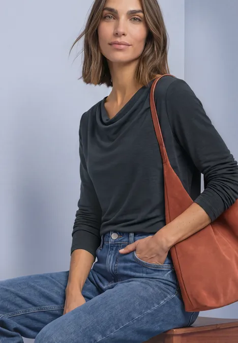 Frau in schwarzem Langarmshirt mit Wasserfallausschnitt, Jeans und brauner Schultertasche sitzt und lächelt.