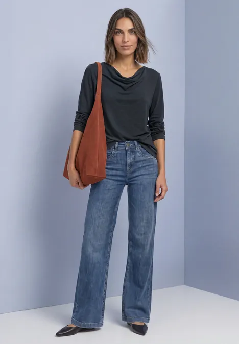Frau vor blauem Hintergrund mit schwarzem Oberteil, weiten Jeans, schwarzen Schuhen und großer brauner Tasche.