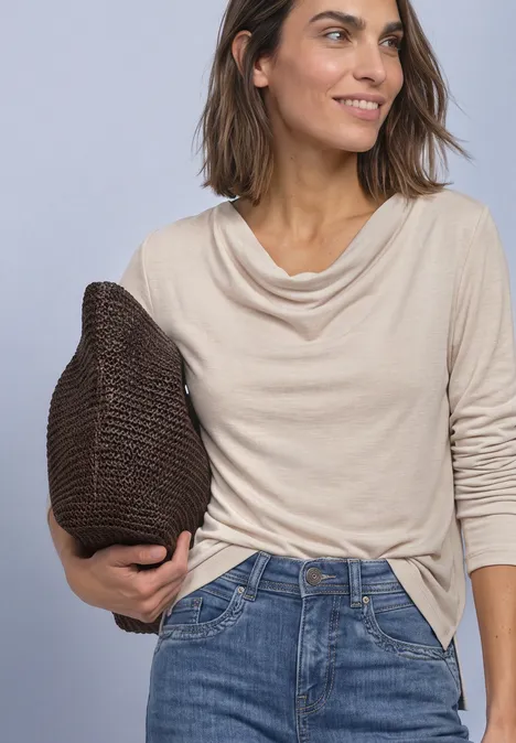 Eine Frau in beigem Oberteil mit Wasserfallausschnitt, Jeans und brauner Stricktasche, lächelnd.