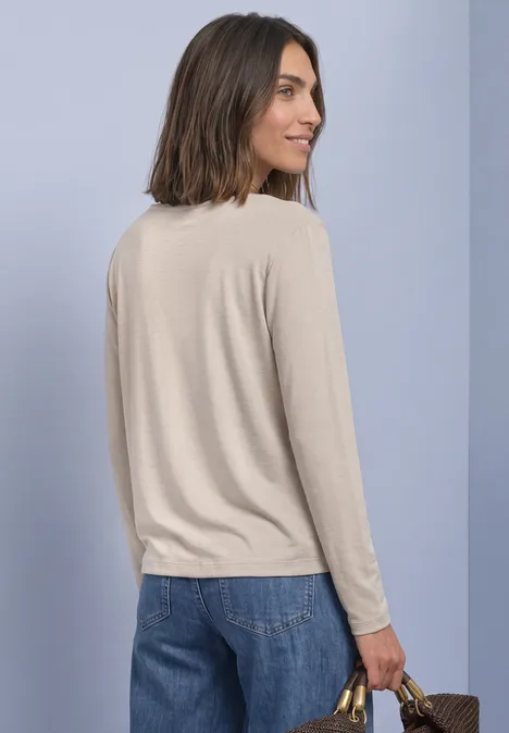 Frau im beigen Longsleeve und Jeans, seitlich stehend, hält eine Handtasche vor hellem Hintergrund.