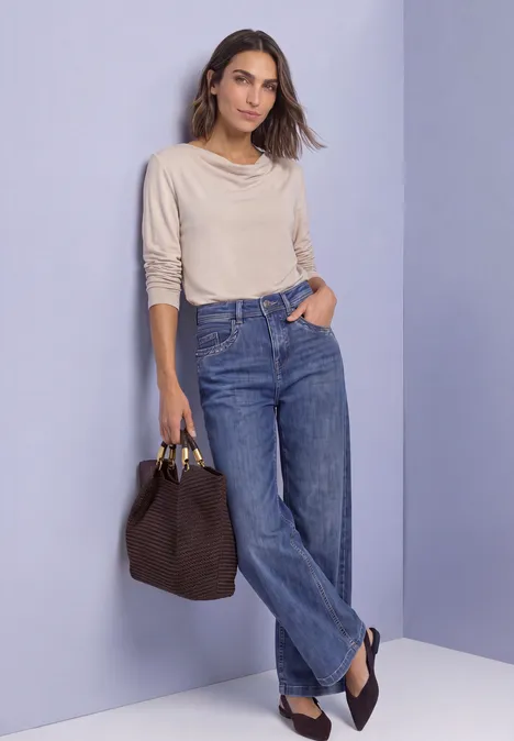Frau in hellem Pullover und weiten Blue-Jeans mit Tasche in der Hand steht locker vor lila Hintergrund.