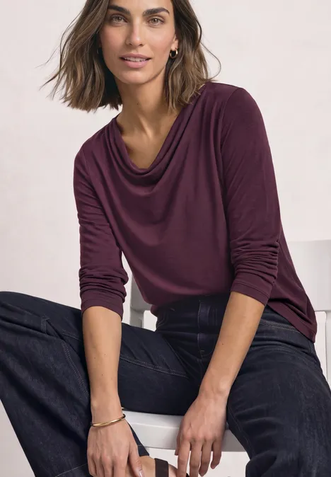 Frau in bordeauxrotem Langarmshirt und dunkler Jeans sitzt lässig, Hände locker auf den Knien.