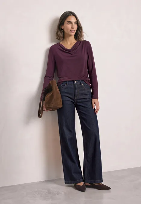 Frau trägt lila Wasserfall-Pullover, weite Jeans, braune Tasche und flache Loafer vor schlichter Wand.