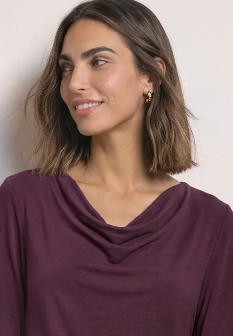 Eine lächelnde Frau mit schulterlangem braunem Haar in einem violetten, drapierten Oberteil, leicht seitlich blickend.