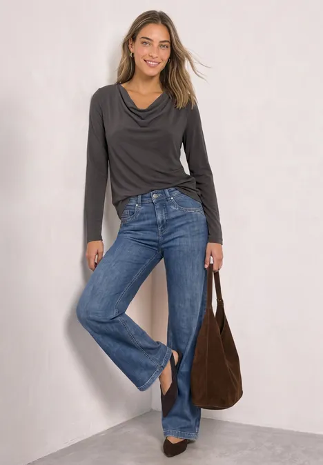 Frau trägt graues Langarmshirt, blaue Jeans und hält eine braune Velourstasche.