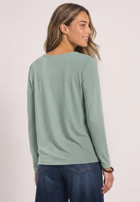 Seitenansicht einer Frau in hellgrünem Langarmshirt und blauen Jeans.