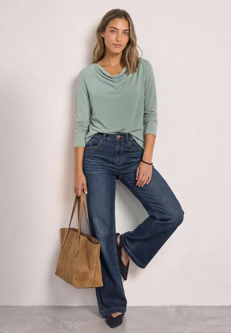 Frau in grünem Wasserfall-Shirt und weit geschnittenen Jeans, mit brauner Wildledertasche und Stiefeletten.