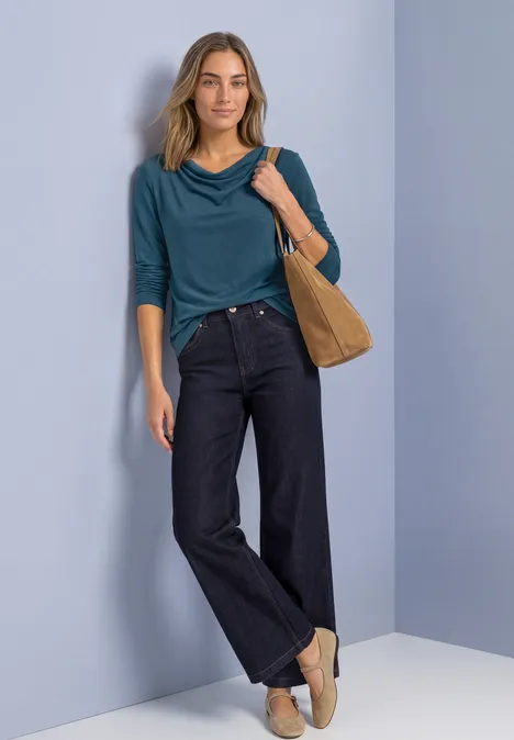 Frau trägt tealfarbenes 3/4-Armshirt, weite dunkle Jeans, beige Slipper und Schultertasche vor hellem Hintergrund.