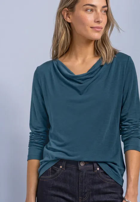 Frau mit blaugrünem Top mit Wasserfallausschnitt und dunkler Jeans blickt zur Seite