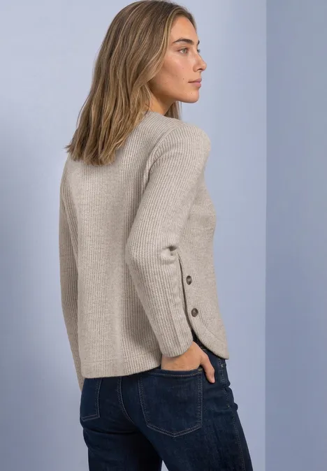 Vrouw in profiel met een beige trui met knoopdetails aan mouwen en een spijkerbroek.