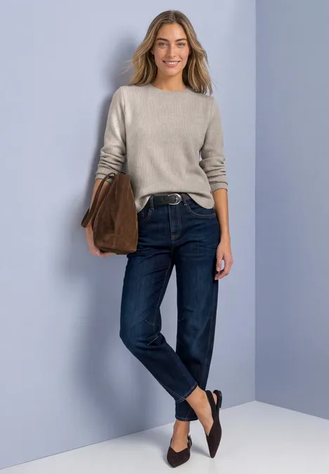 Een vrouw draagt een trui, donkere jeans, zwarte ballerinas en houdt een tas vast.