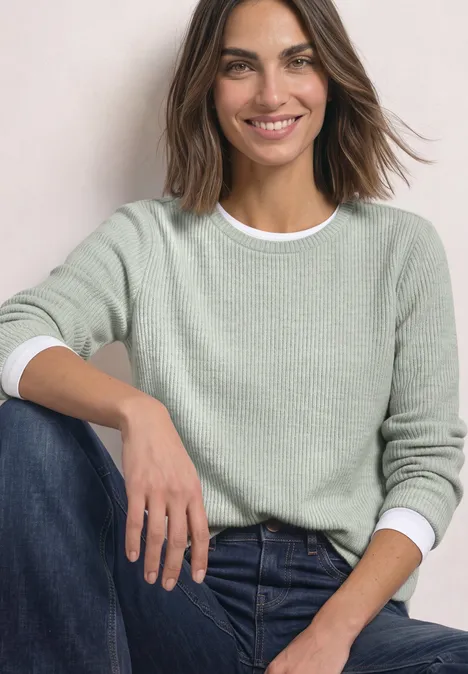 Een glimlachende vrouw draagt een lichtgroene trui en blauwe jeans, zittend voor een egale achtergrond.